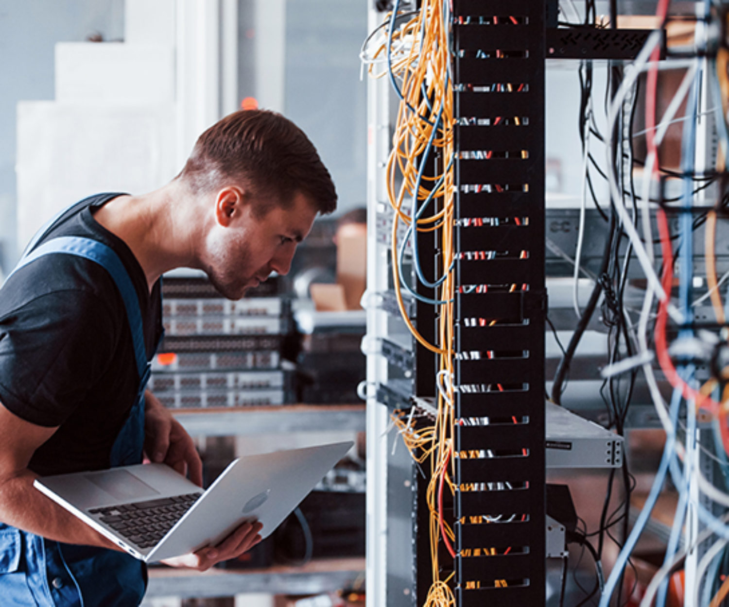 Young man in uniform and with laptop works with internet equipment and wires in server room.